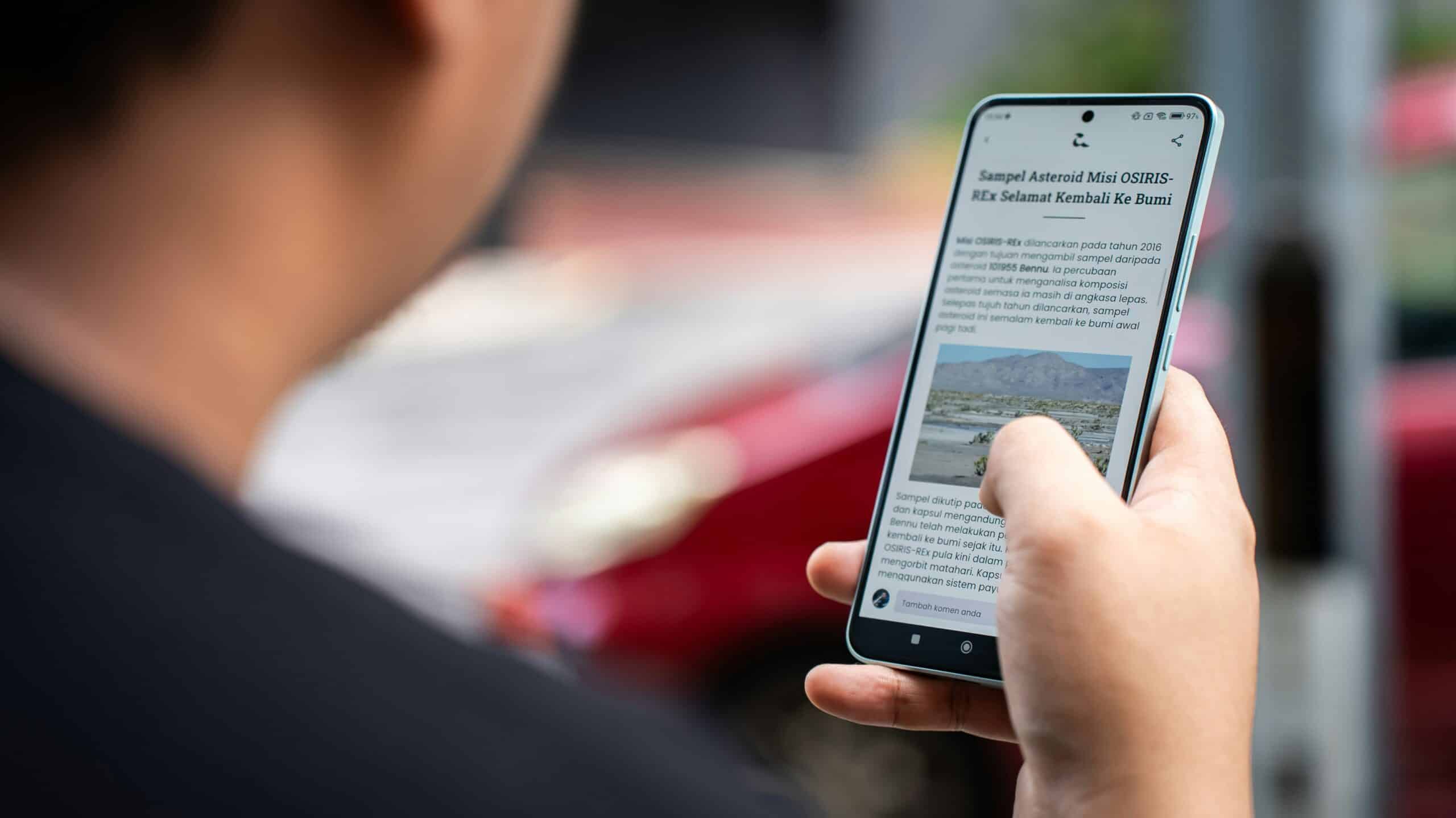A person uses their smartphone to read an article, pressing the screen to enlarge a photo of a landscape with text in a foreign language. The individual is holding the phone in their left hand and is outdoors. The background is blurred but shows a red car.
