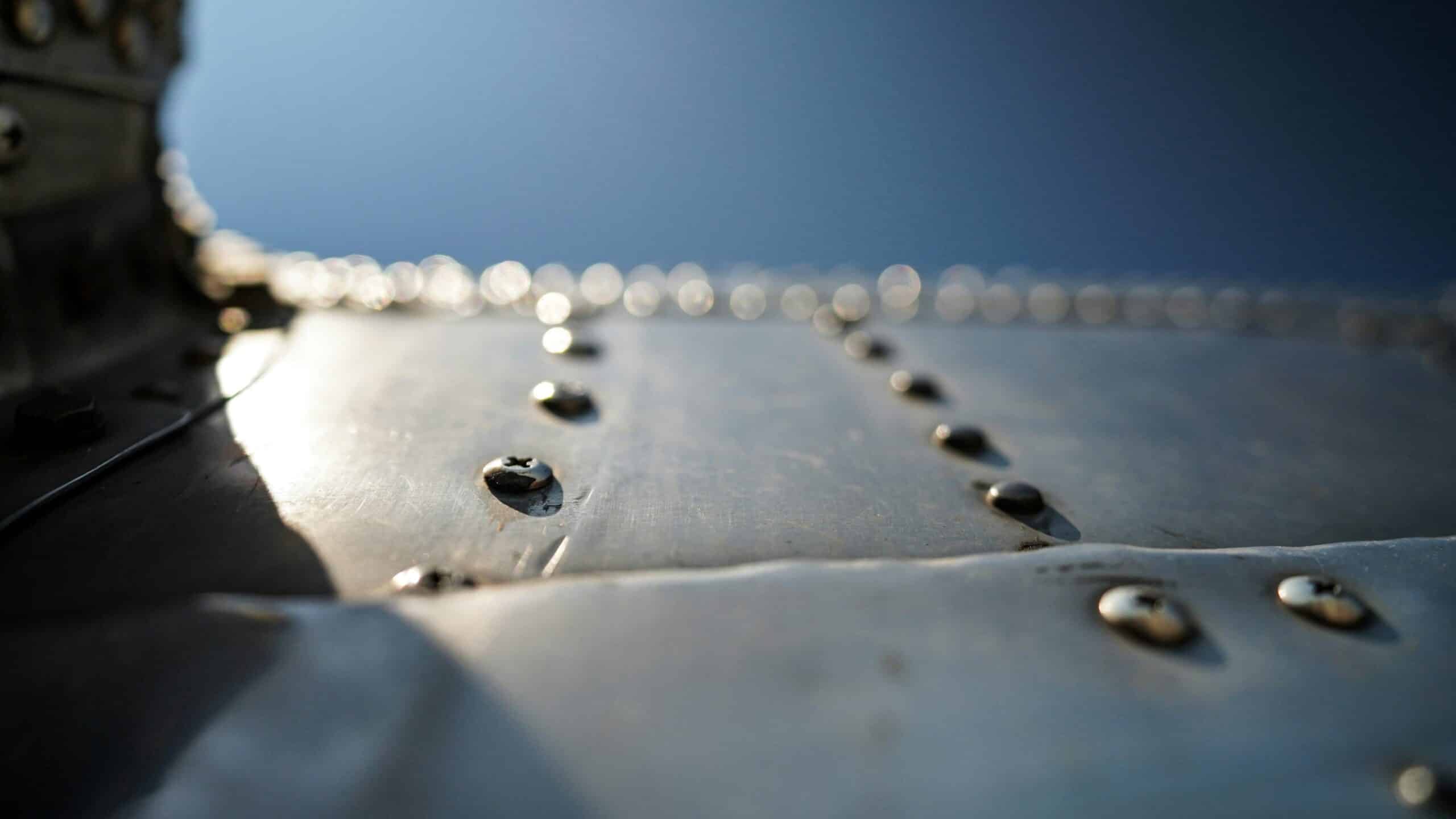 Close-up of an aluminium surface with rivets against a blurred blue background. The sunlight creates a soft glow on the polished metal, showcasing the texture and craftsmanship of the riveted panels. The image exudes an industrial and rugged aesthetic, reminiscent of a transport gallery exhibit.