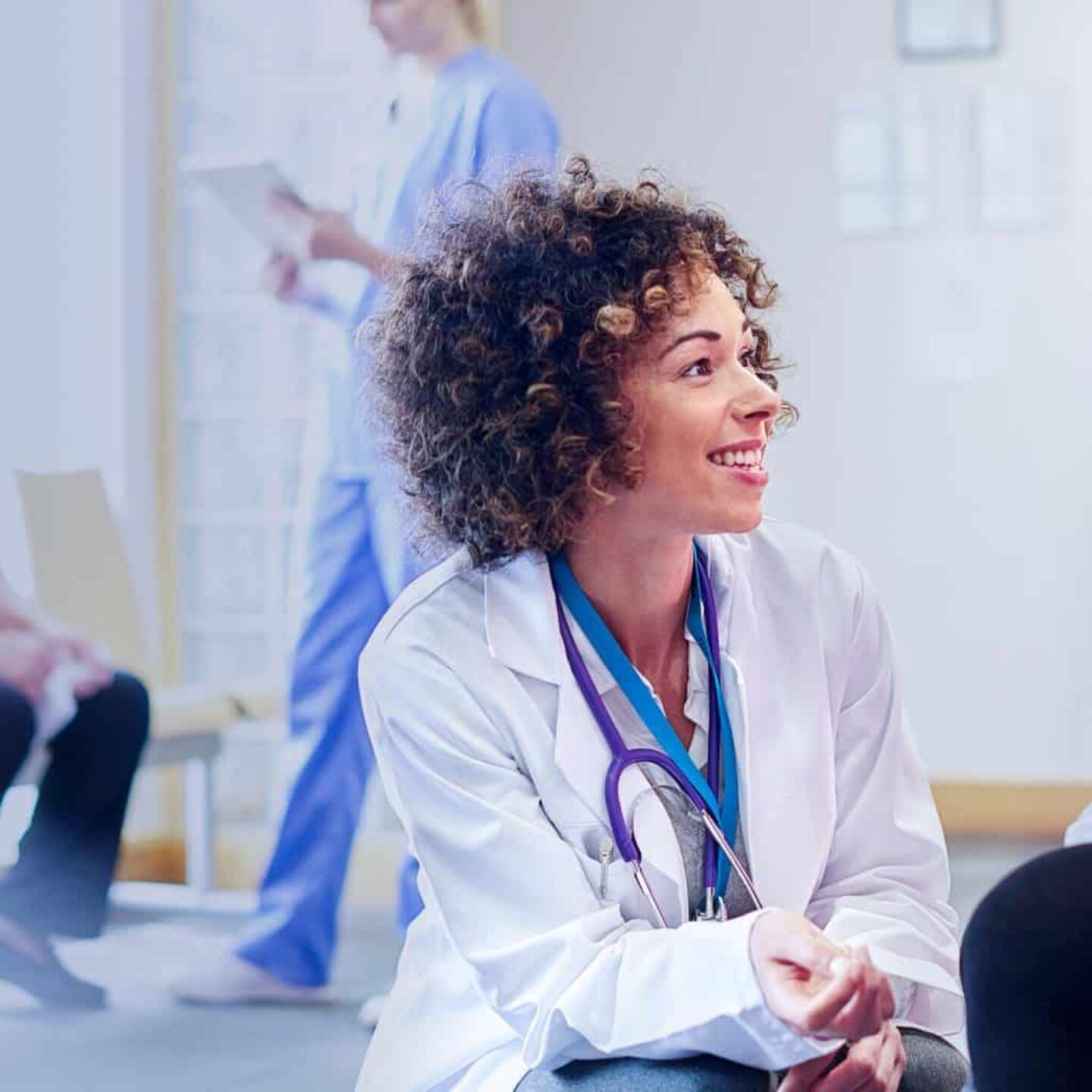 A doctor with curly hair and a stethoscope kneels while talking to a young child wearing a yellow shirt who is sitting on an adult's lap. In the brightly lit clinic setting of the International Aluminium Institute, other medical staff and patients are visible in the background.