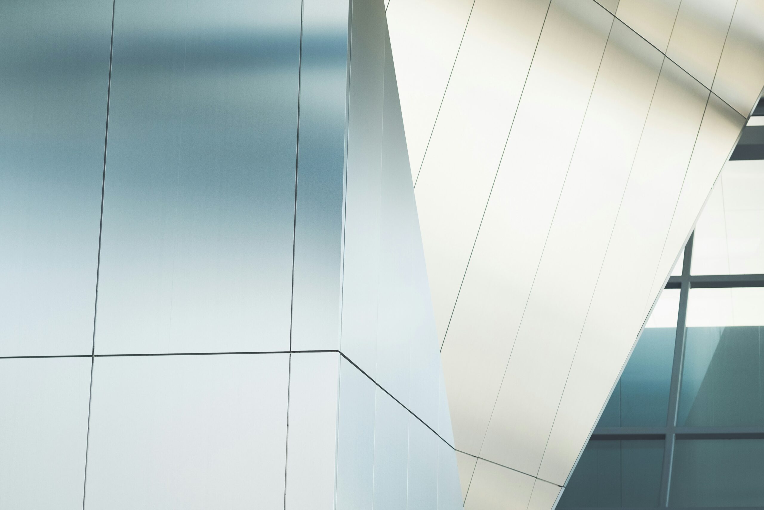 Close-up of a modern building exterior featuring smooth, light-colored aluminium panels. The structure exhibits sharp, angular lines and geometric shapes, reminiscent of contemporary gallery buildings. One section reflects light more brightly, indicating a slight bend or fold in the surface. The overall look is sleek and minimalist.