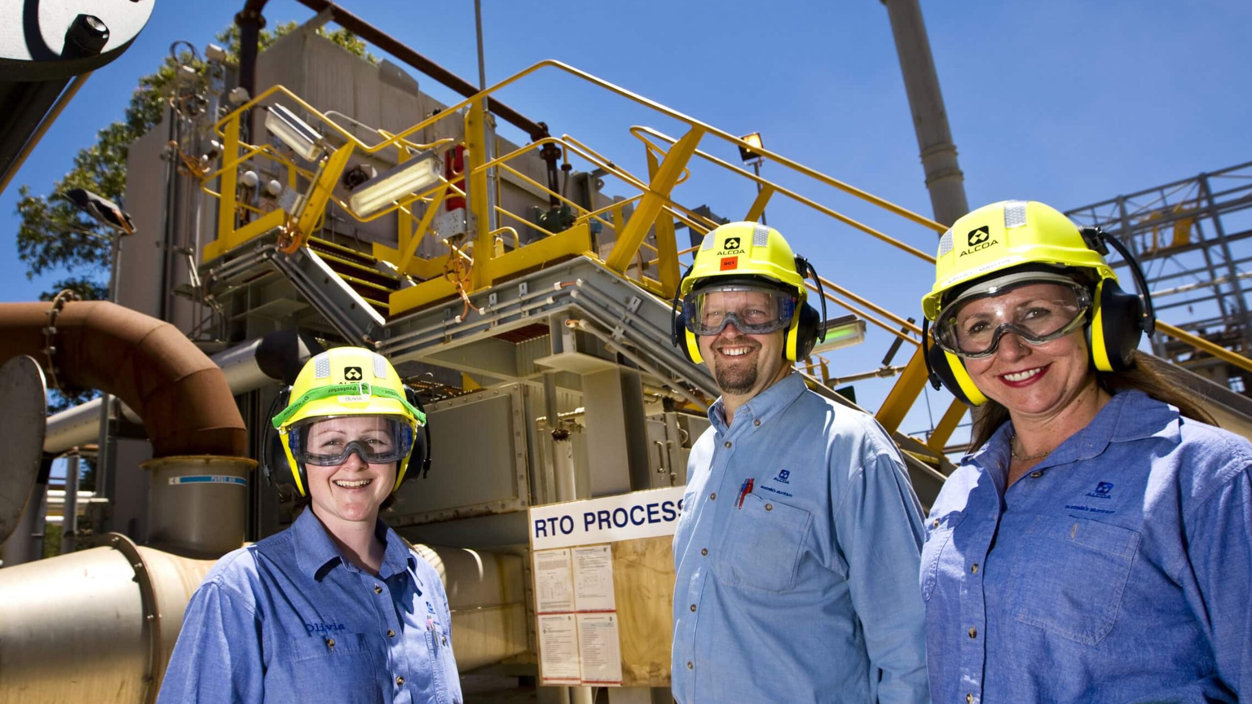 Three people wearing yellow helmets, goggles, and blue shirts stand in front of industrial equipment on a sunny day. Emphasizing occupational health, they all wear ear protection and look towards the camera, smiling. A sign behind them reads "RTO PROCESS," with metal structures and aluminium pipes visible in the background.