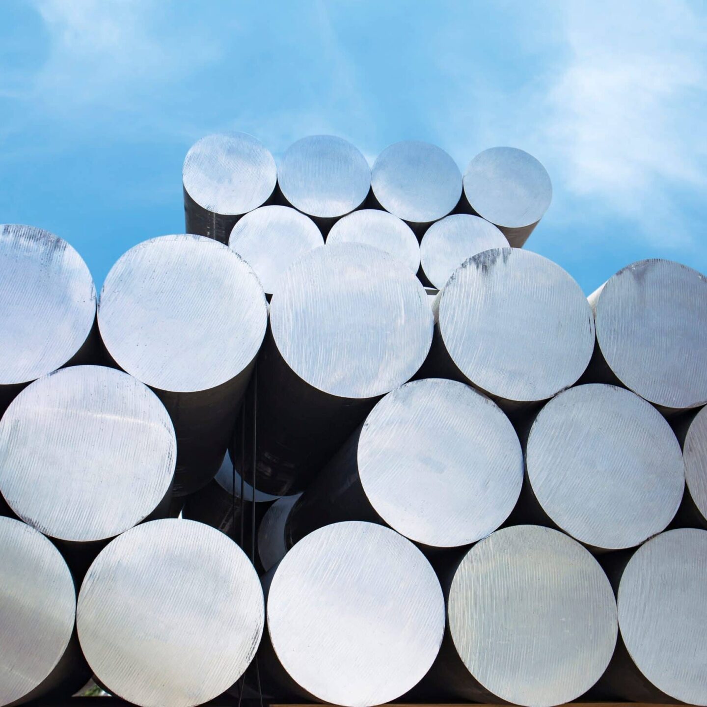 A stack of large, silver metallic cylinders is arranged in neat rows against a clear blue sky. The aluminium cylinders reflect light, showcasing their smooth, polished surfaces. The perspective emphasizes the uniformity and industrial nature of the objects, capturing 50 moments of precision.