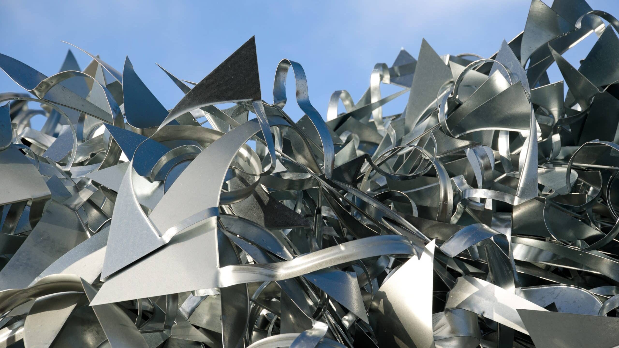 A pile of sharp, angular aluminium scrap with various shapes and edges is heaped against a backdrop of a clear blue sky. The metallic pieces reflect light, creating a shiny, contrasting surface. A pile of sharp, angular aluminium scrap with various shapes and edges is heaped against a backdrop of a clear blue sky. The metallic pieces reflect light, creating a shiny, contrasting surface.