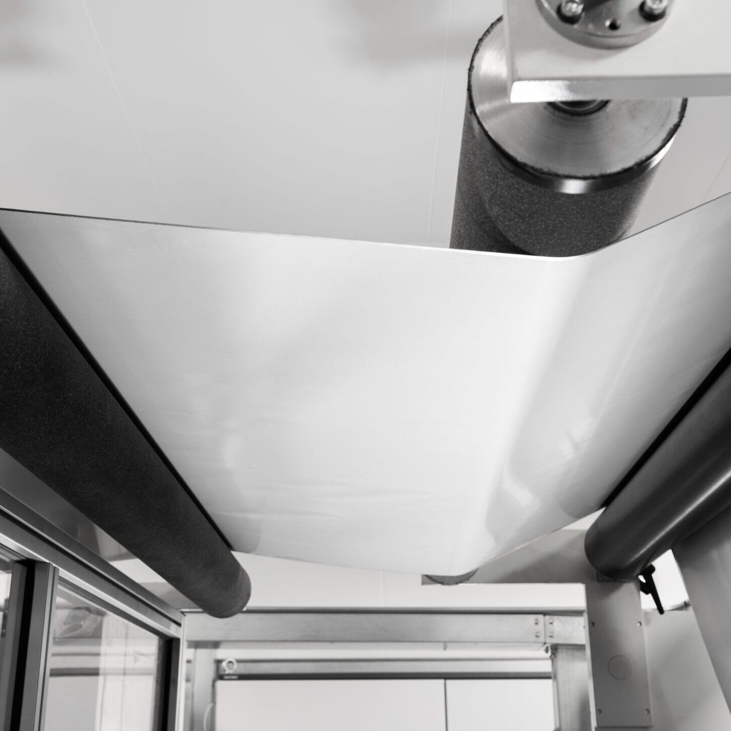 A sheet of white material being processed through an industrial roller machine with sleek aluminium parts and black padded rollers, seen in monochrome tones, suggesting a manufacturing or production setting that feels futuristic, perhaps hinting at the advancements of 2030.