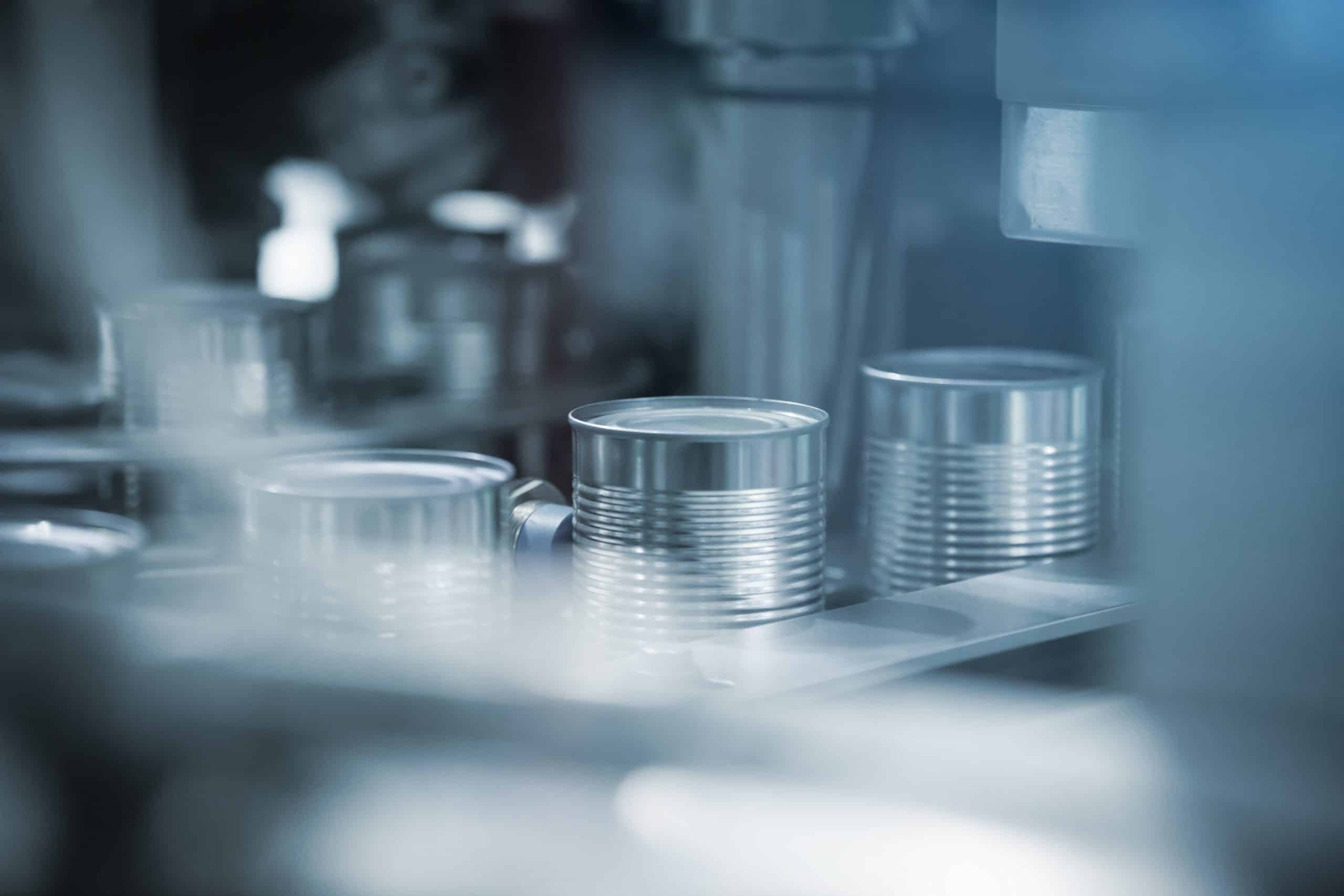 A close-up shot of a commercial canning line in a factory. Several silver tin cans are visible, positioned in a row on a moving conveyor belt, surrounded by blurred machinery components in the foreground and background, highlighting the process that significantly impacts carbon footprints.