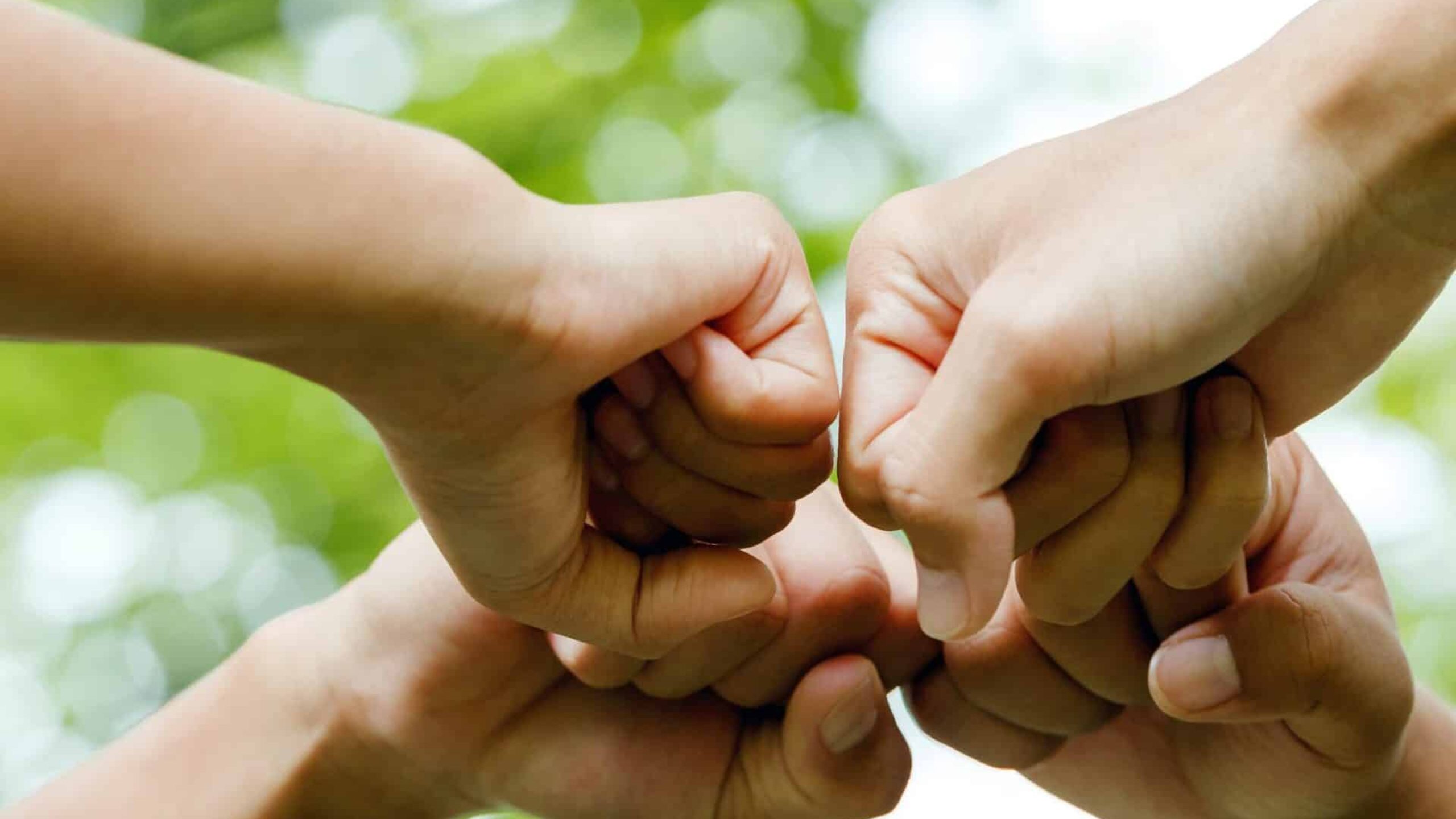 A close-up image of six hands engaged in a fist bump, forming a circle against a blurred green background. The hands are of different individuals coming together, symbolizing unity, teamwork, and the strength of communities. The natural greenery in the background suggests an outdoor setting.