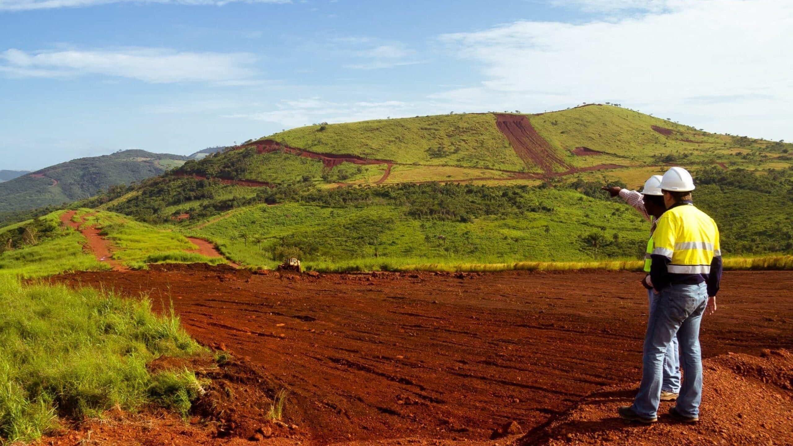 Two workers, clad in yellow and white safety gear and helmets, stand on red dirt ground. One points towards a green, hilly landscape with dirt tracks under a blue sky scattered with clouds. The area, likely a construction or mining site, may be exploring potential bauxite residue use.