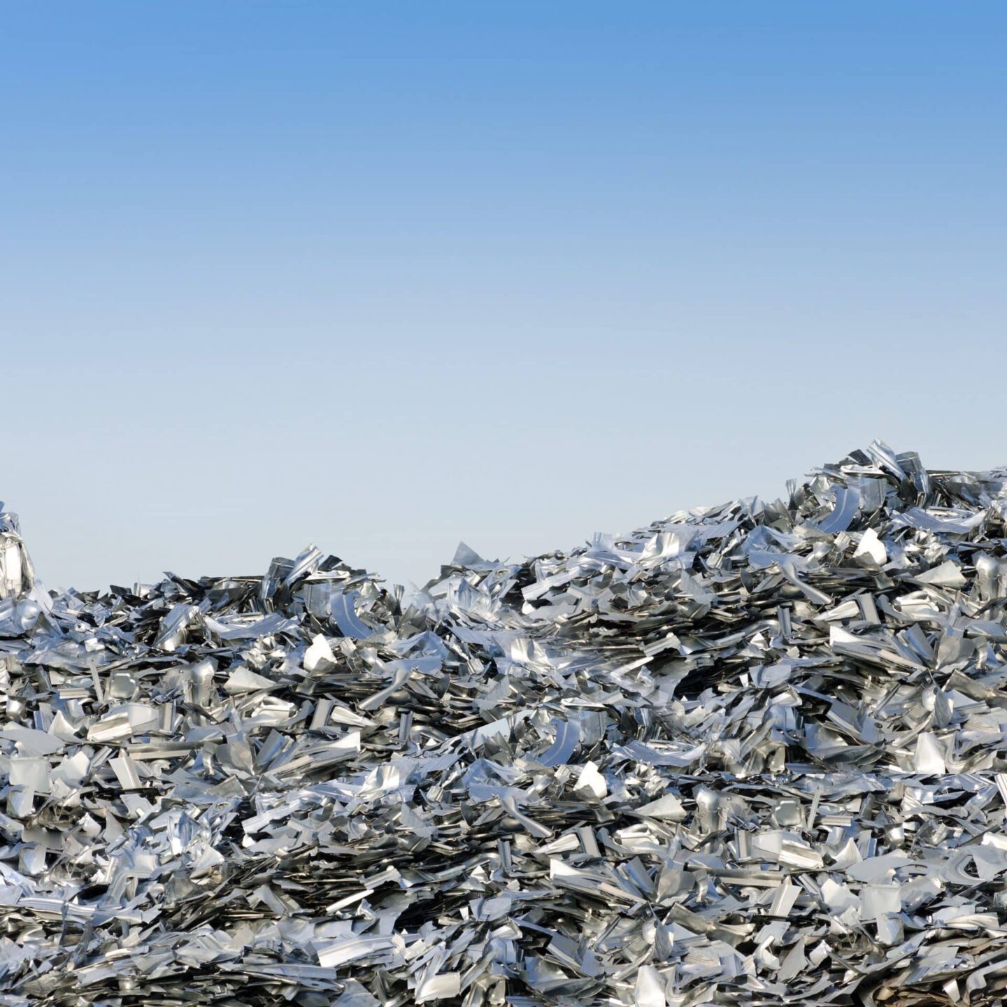 A vast pile of crushed and flattened metallic scrap pieces, primarily for recycling aluminium, stretches into the horizon under a clear blue sky. The metal shards reflect light, creating a shimmering effect across the heap of debris.