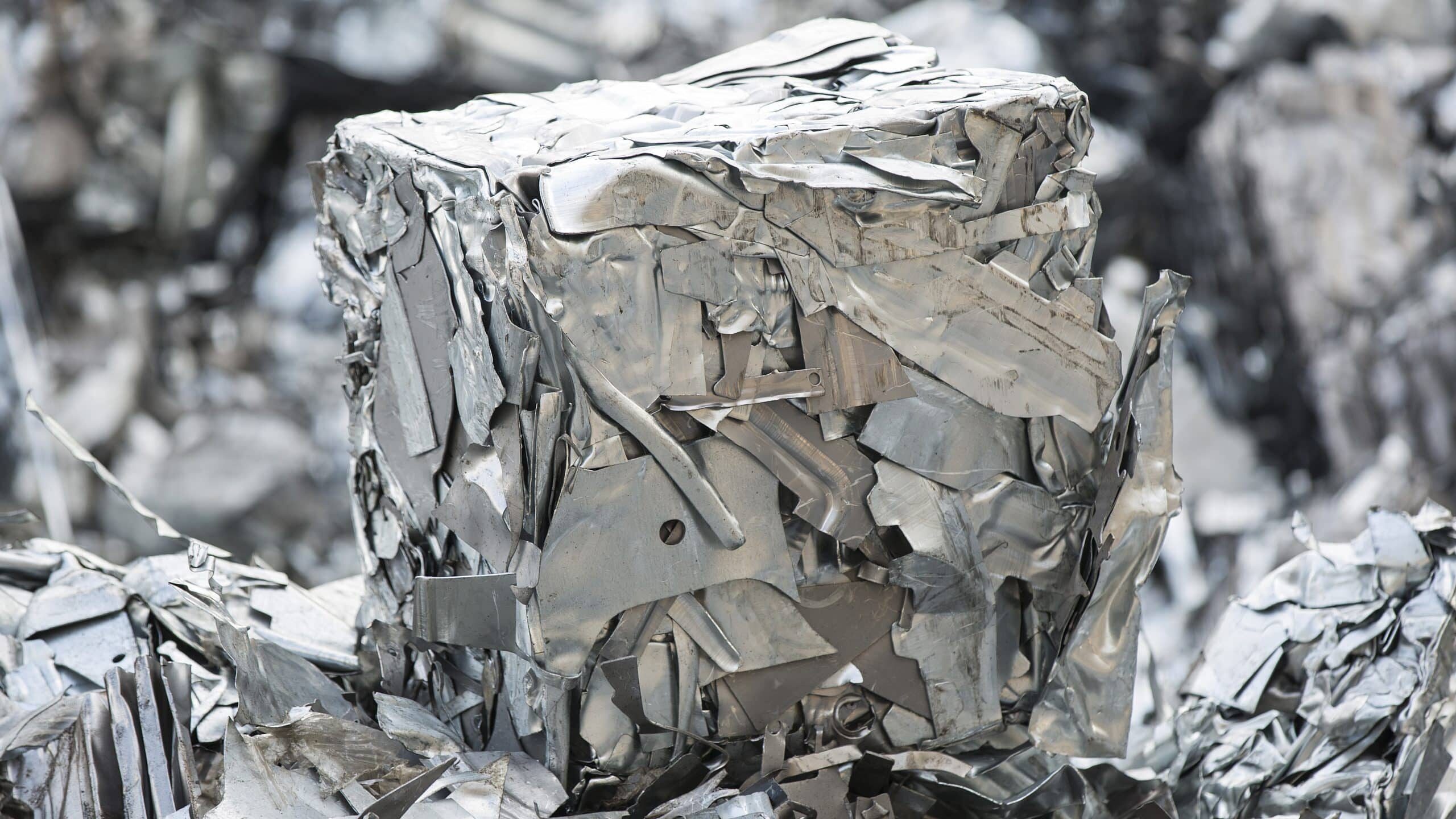A compacted cube of crushed and flattened scrap metal, primarily made of aluminium, surrounded by more scrap metal in various shapes and sizes. The pieces appear silver and metallic, with rough and jagged edges, reflecting light from their surfaces. This scene could be essential for a detailed material flow analysis.