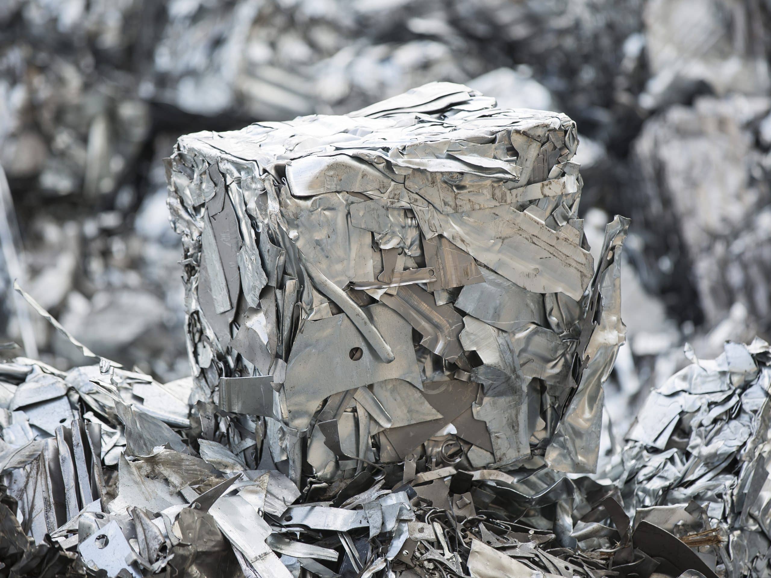A compacted cube of crushed and flattened scrap metal, primarily made of aluminium, surrounded by more scrap metal in various shapes and sizes. The pieces appear silver and metallic, with rough and jagged edges, reflecting light from their surfaces. This scene could be essential for a detailed material flow analysis.